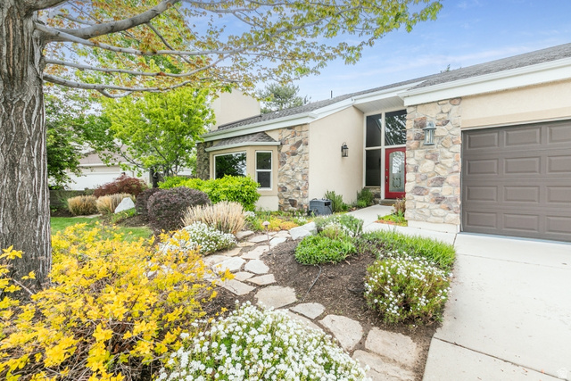 10183 Hickory Point Drive Sandy, UT 84092 - Photo 4 of 36 Exterior featuring a stone and stucco facade, a prominent red entry door, an attached garage with a panel door, a bay window, and a flagstone pathway surrounded by diverse plantings