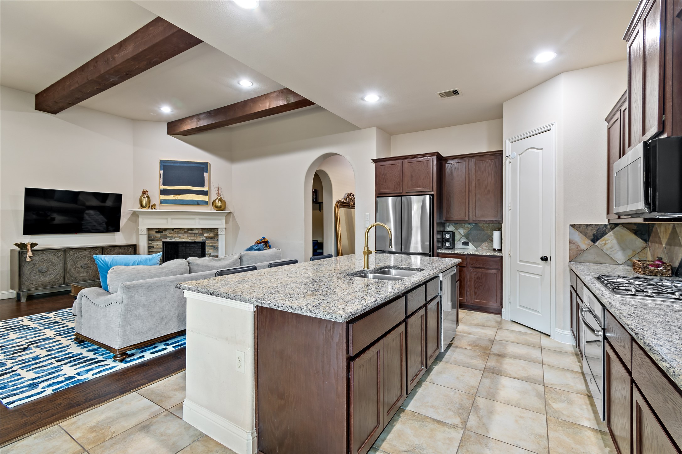 8126 Spreadwing Street Conroe, TX 77385 - Photo 13 of 30 This photo showcases a spacious open-concept kitchen and living area. The kitchen features a large island with granite countertops, stainless steel appliances, and dark wood cabinetry. The living area includes a cozy gas fireplace and space for seating, complemented by exposed ceiling beams for added character.