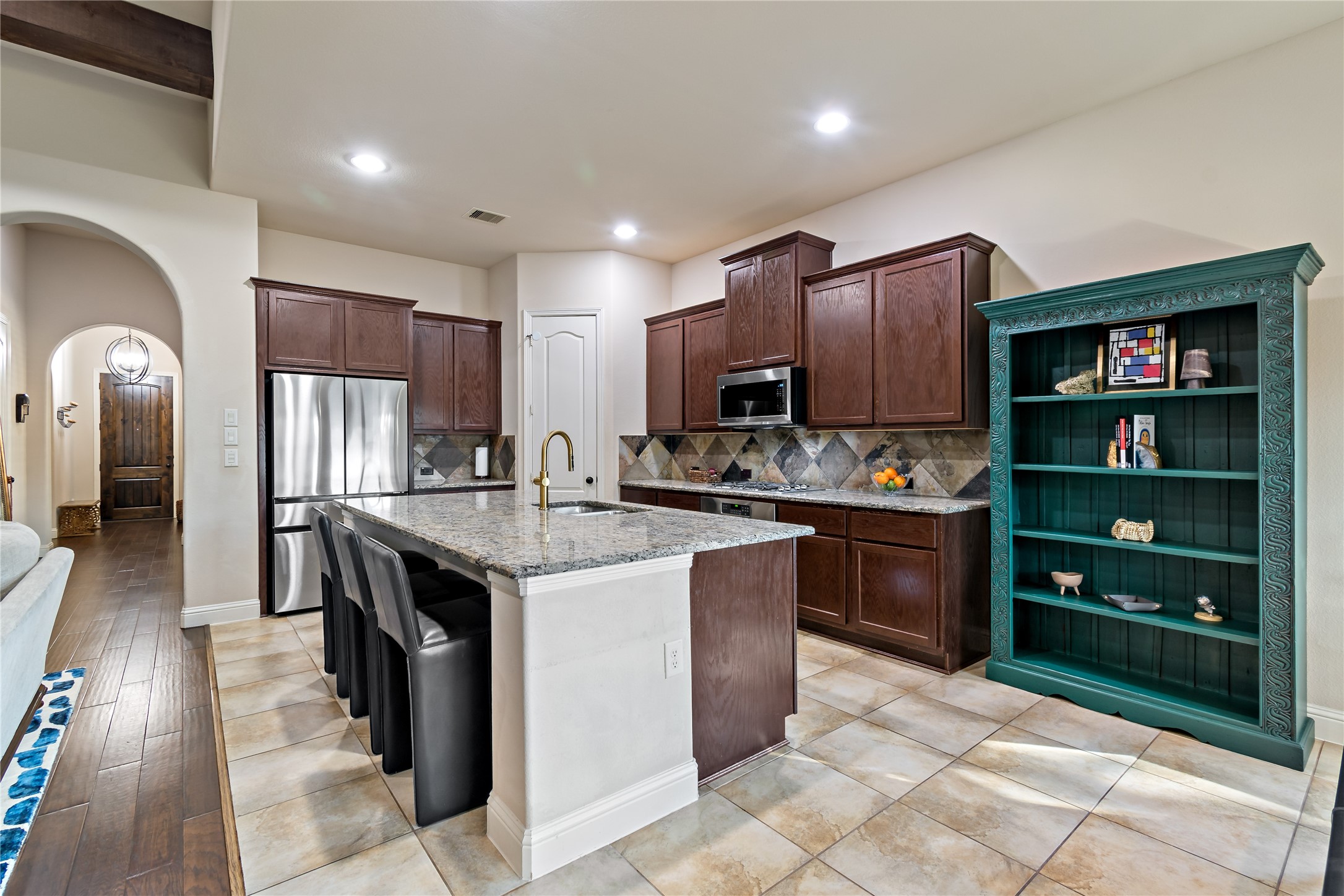 8126 Spreadwing Street Conroe, TX 77385 - Photo 18 of 30 This kitchen features dark wood cabinets, a large island with granite countertops and seating, stainless steel appliances, and a decorative slate backsplash. The space is bright with recessed lighting and opens to a hallway with wood flooring. A colorful shelving unit adds a touch of personality.