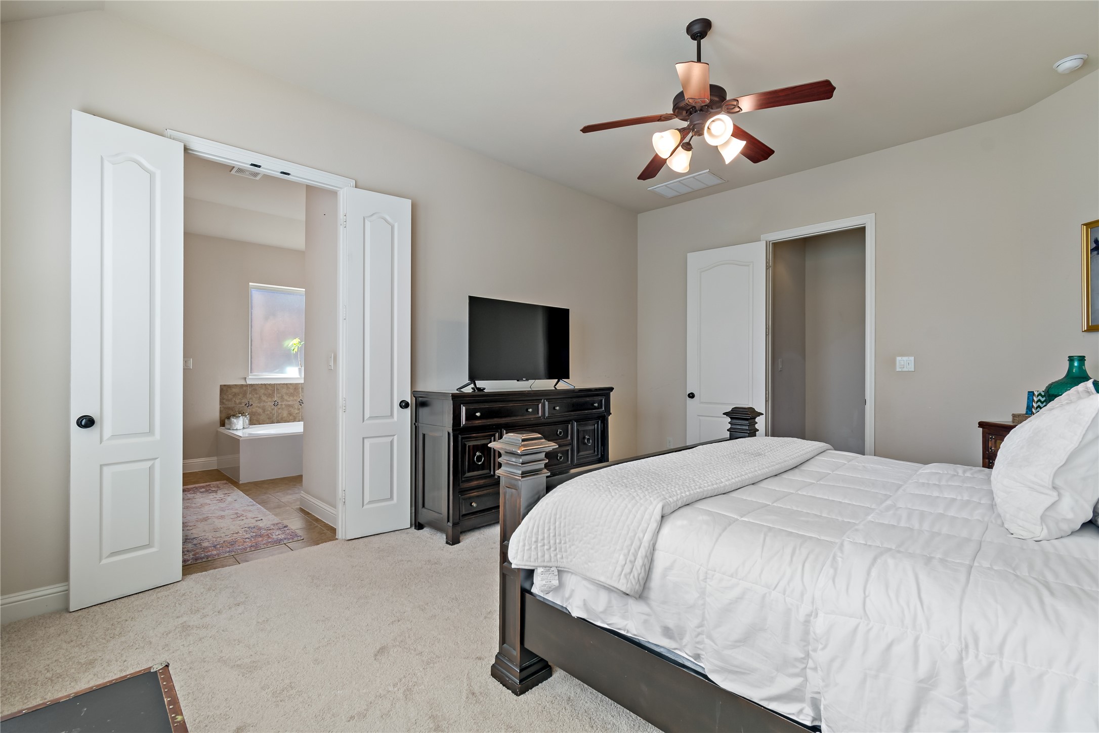 8126 Spreadwing Street Conroe, TX 77385 - Photo 19 of 30 This primary bedroom shows a spacious bedroom with a neutral color scheme, featuring a ceiling fan, a large bed, and a wall-mounted TV. There is an en-suite bathroom visible through double doors, adding convenience and luxury to the space.