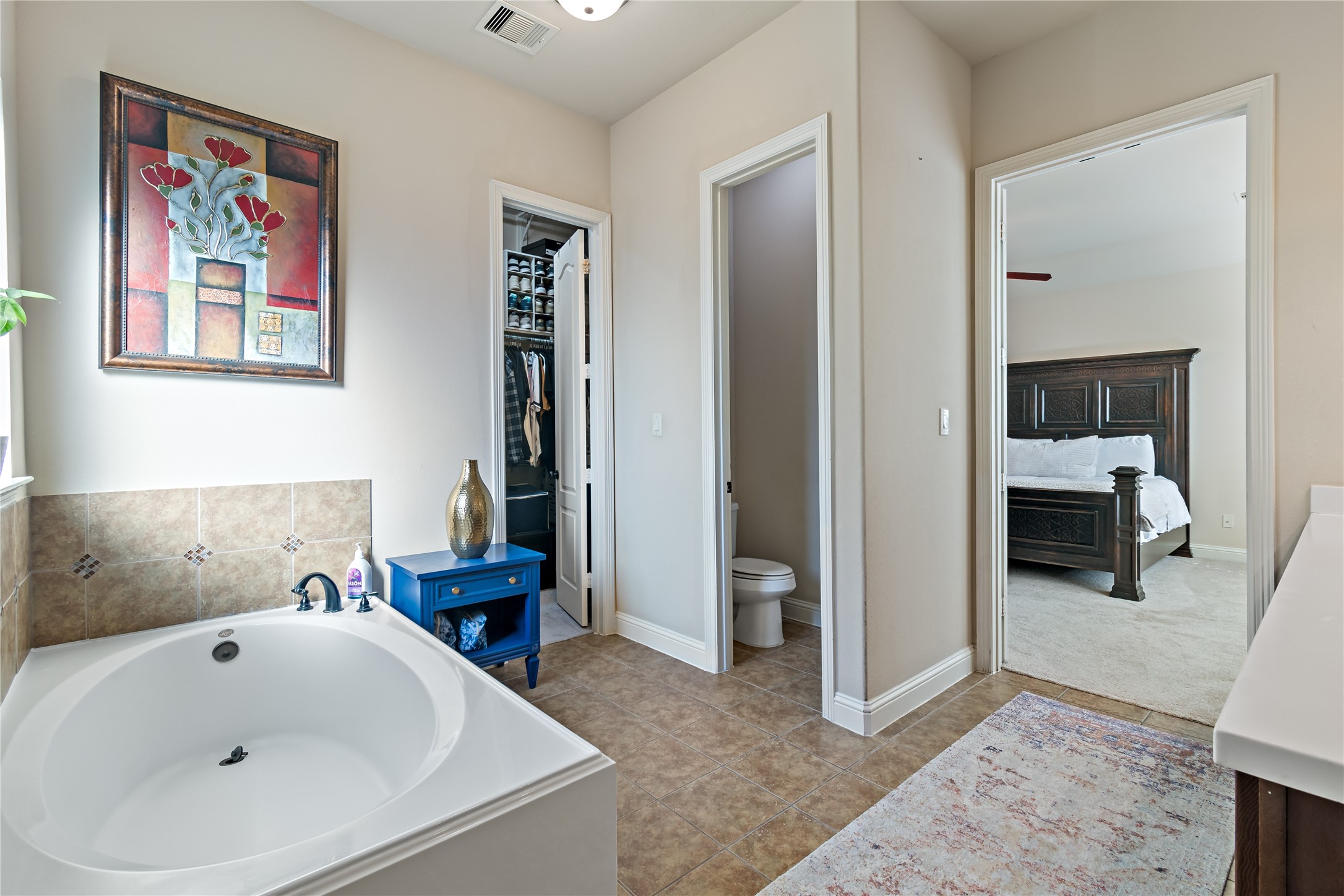 8126 Spreadwing Street Conroe, TX 77385 - Photo 21 of 30 This primary bathroom shows a bright bathroom with a large soaking tub, decorative wall art, and a view into an adjacent bedroom. The space includes a small blue cabinet and neutral tones, creating a cozy and inviting atmosphere.