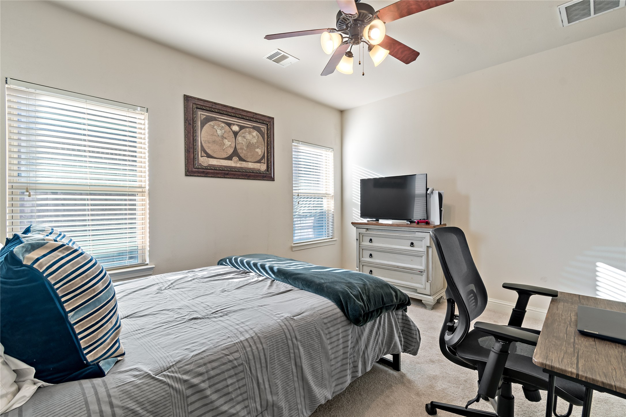 8126 Spreadwing Street Conroe, TX 77385 - Photo 24 of 30 This is a cozy bedroom featuring a bed with striped bedding, a ceiling fan, and ample natural light from two windows. It includes a small workspace with a desk and chair, and a dresser with a TV on top.