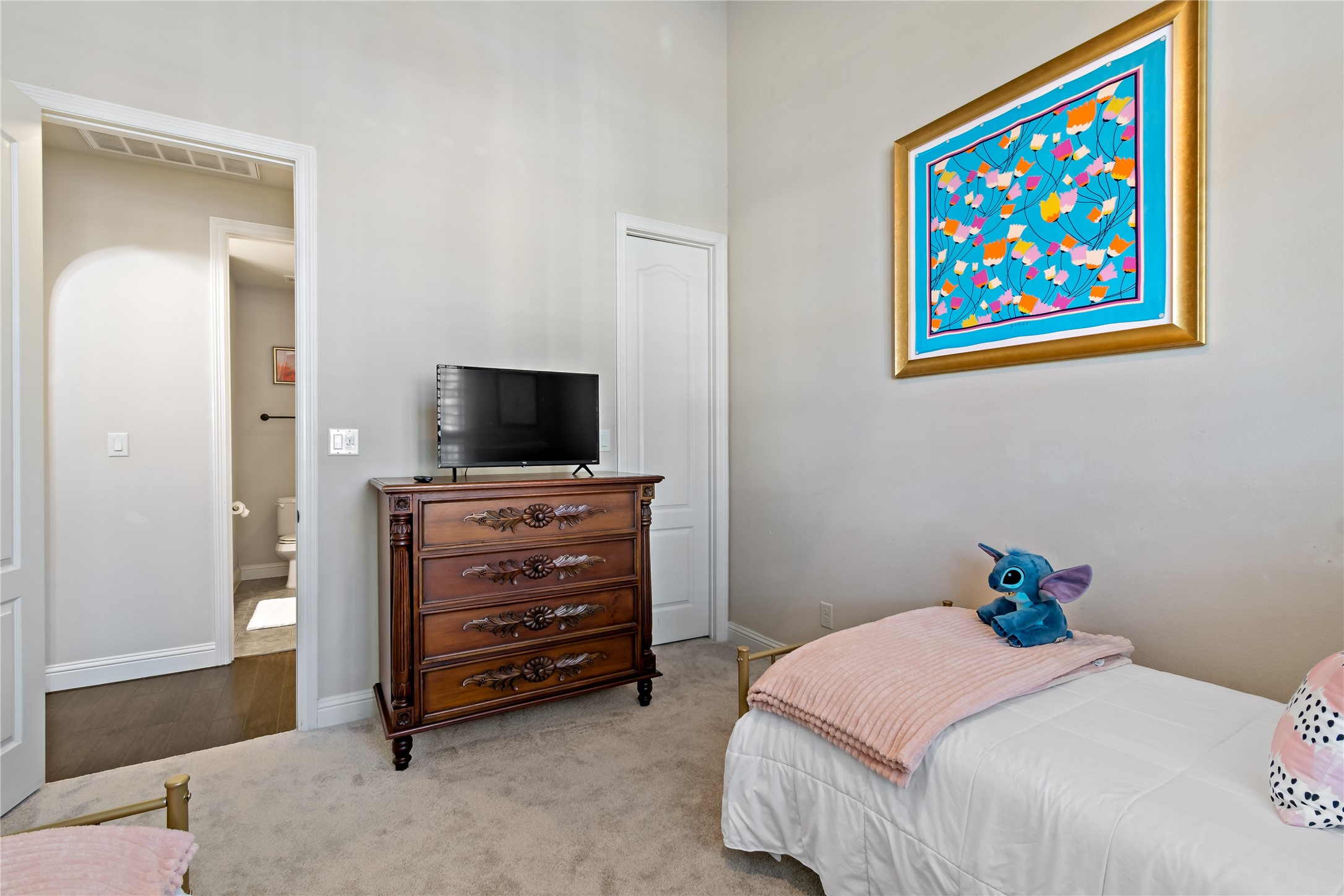 8126 Spreadwing Street Conroe, TX 77385 - Photo 28 of 30 Cozy bedroom featuring a plush carpet, twin bed with pink accents, and a whimsical plush toy. The room includes a wooden dresser with a TV and colorful artwork on the wall. There's an en-suite bathroom visible through an open door, enhancing convenience and privacy.