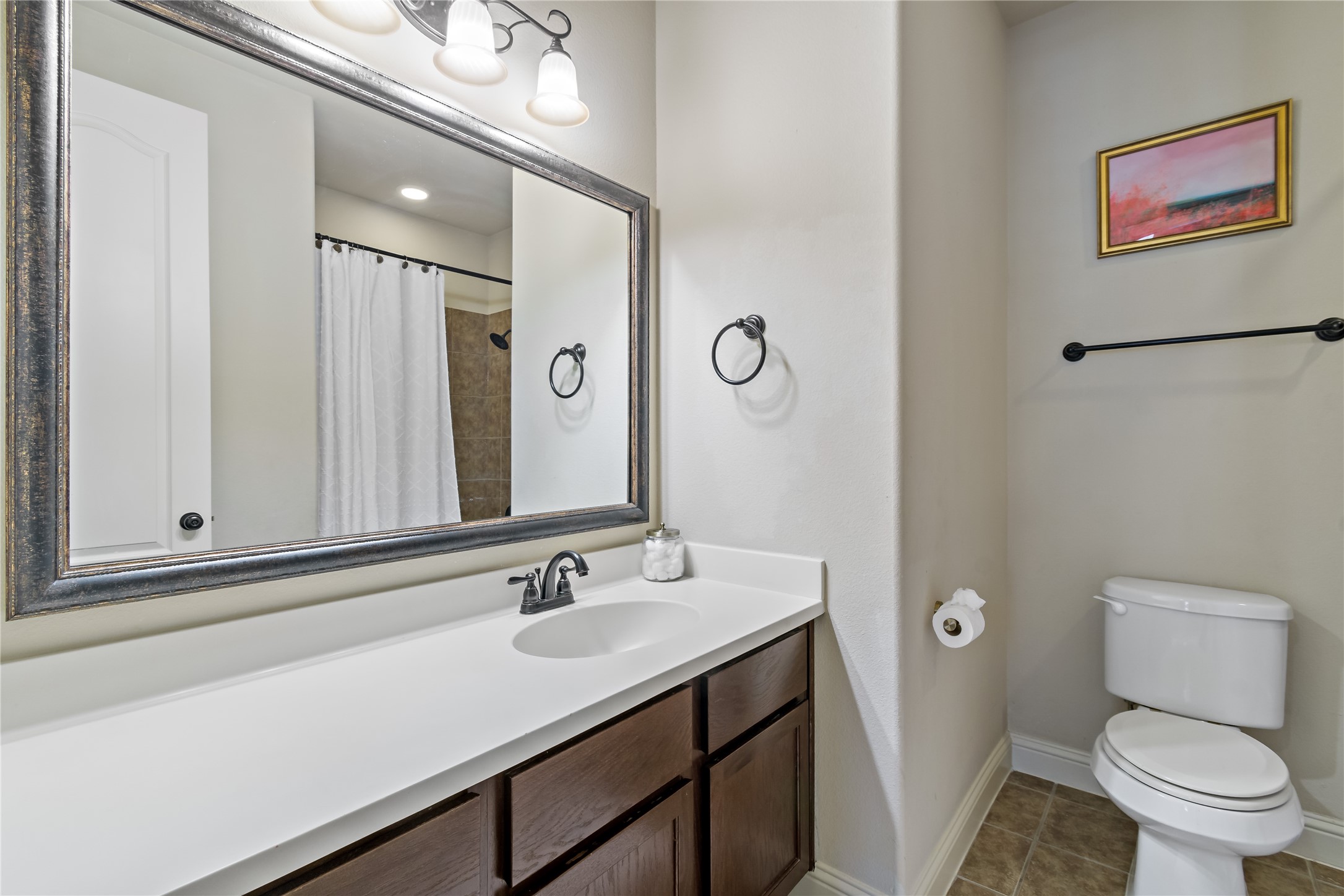 8126 Spreadwing Street Conroe, TX 77385 - Photo 29 of 30 This bathroom features a modern design with a large mirror, a sleek white countertop with built-in sink, and dark wood cabinetry. It includes a shower with a white curtain, a toilet, and a simple wall-mounted towel ring. The space is bright, with neutral tones and a small piece of artwork adding a touch of color.