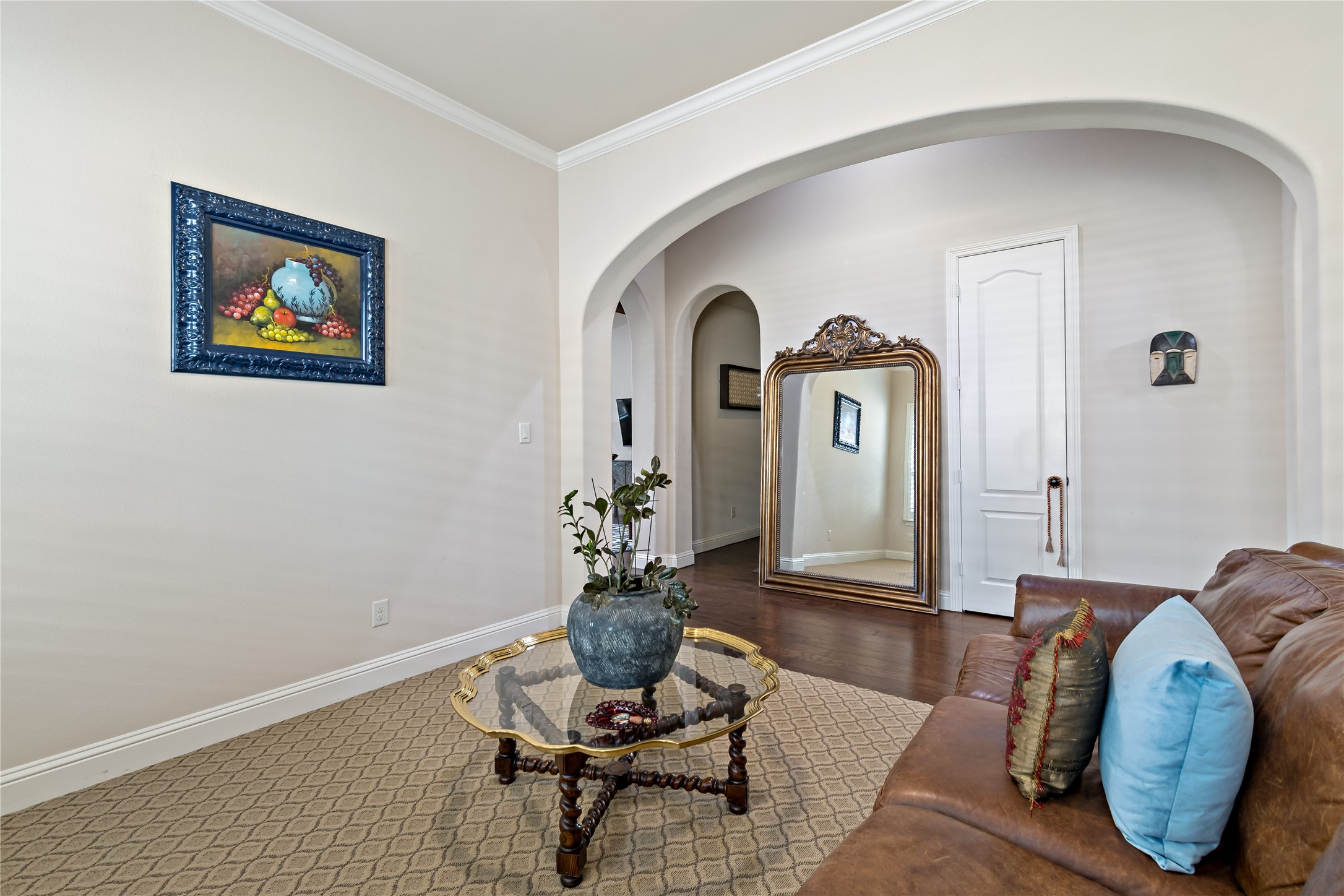 8126 Spreadwing Street Conroe, TX 77385 - Photo 4 of 30 This inviting living area features a cozy brown leather couch, a stylish glass coffee table with a plant centerpiece, and elegant decor. A large mirror and tasteful artwork enhance the room's spacious feel, complemented by a soft, neutral color palette and arched doorway.