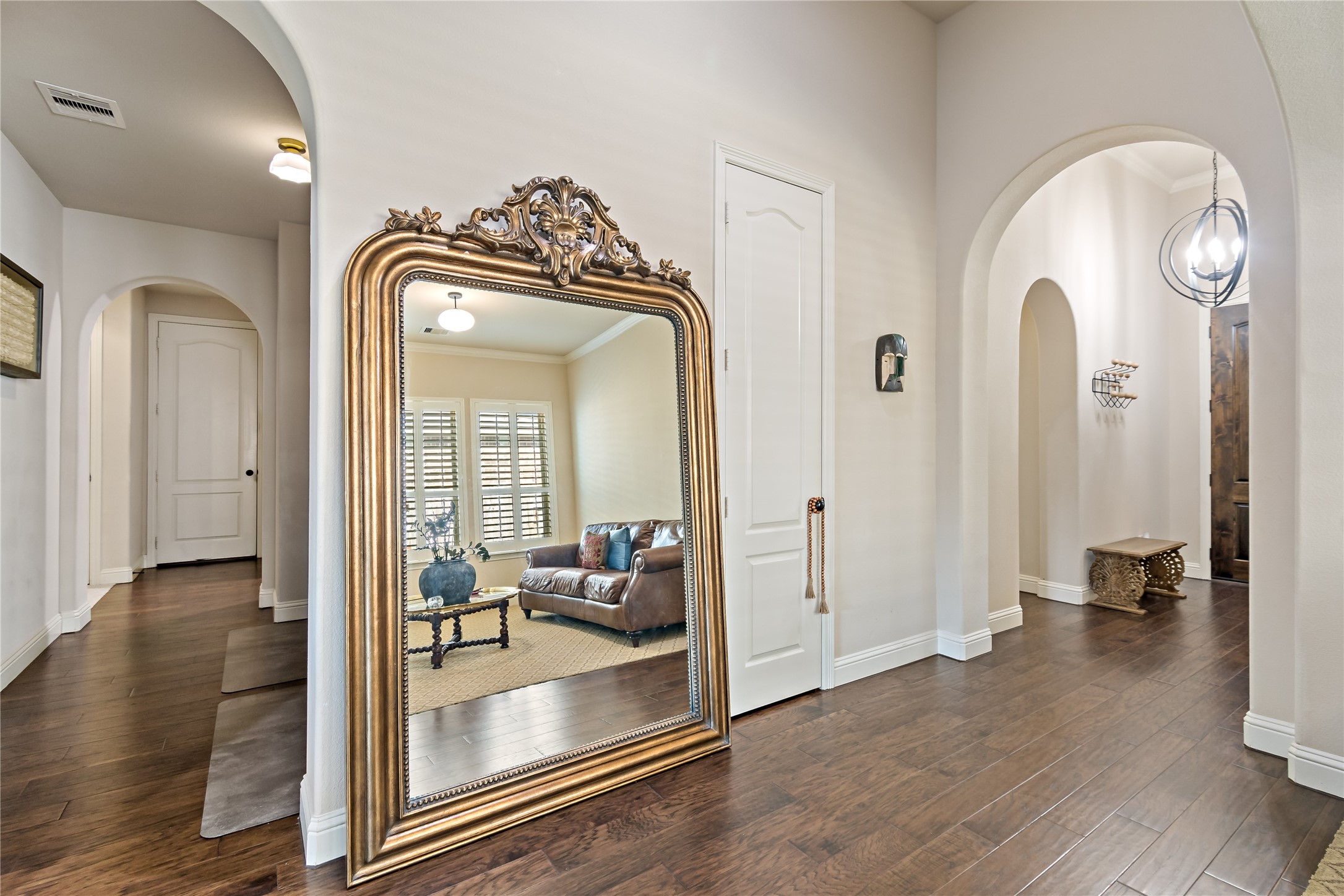 8126 Spreadwing Street Conroe, TX 77385 - Photo 5 of 30 This photo shows a bright, elegant hallway with dark hardwood floors and arched doorways. A large ornate mirror leans against the wall, reflecting a cozy living area with ample natural light. The space feels open and inviting.