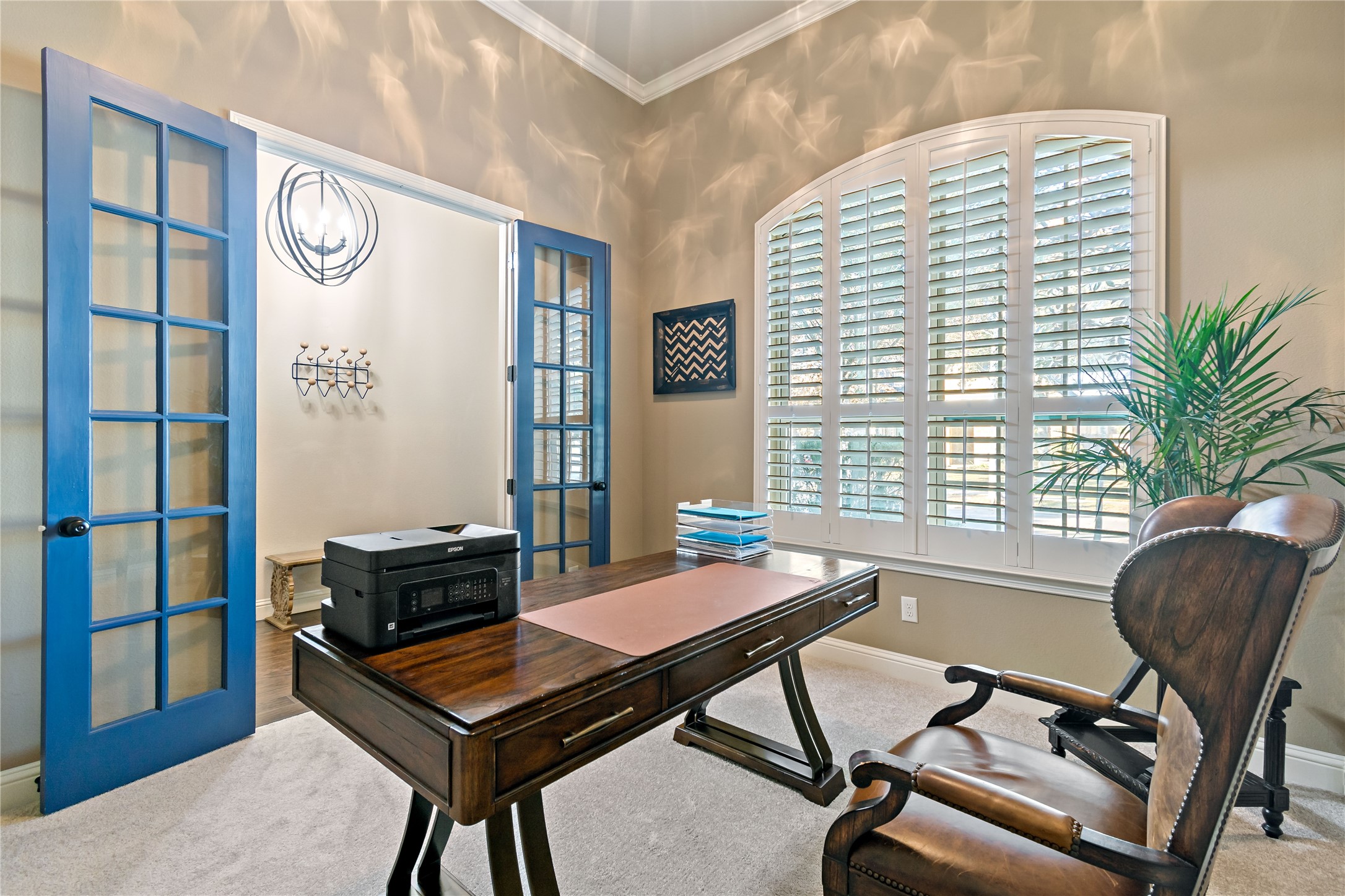 8126 Spreadwing Street Conroe, TX 77385 - Photo 8 of 30 This home office features elegant blue French doors, a large window with plantation shutters for natural light, and a classic wooden desk with a comfortable chair. The space is accentuated with neutral tones and a touch of greenery, creating a bright and inviting work environment.