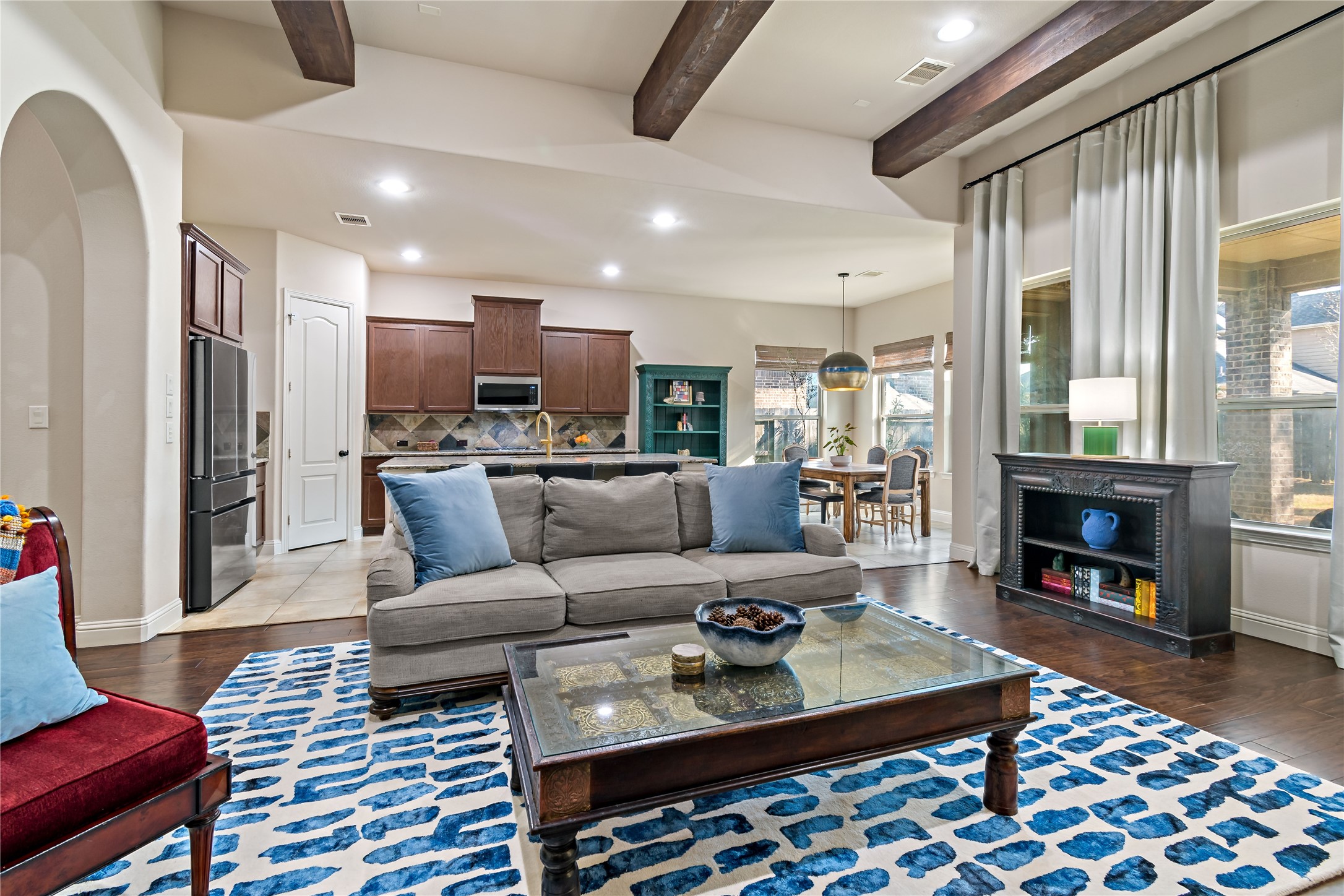 8126 Spreadwing Street Conroe, TX 77385 - Photo 10 of 30 This photo shows a spacious open-concept living area with a cozy sitting space, accented by a blue patterned rug and neutral-toned furniture. The room features exposed wooden beams, recessed lighting, and large windows that allow natural light. The kitchen in the background has modern cabinetry and stainless-steel appliances, creating an inviting and functional space.