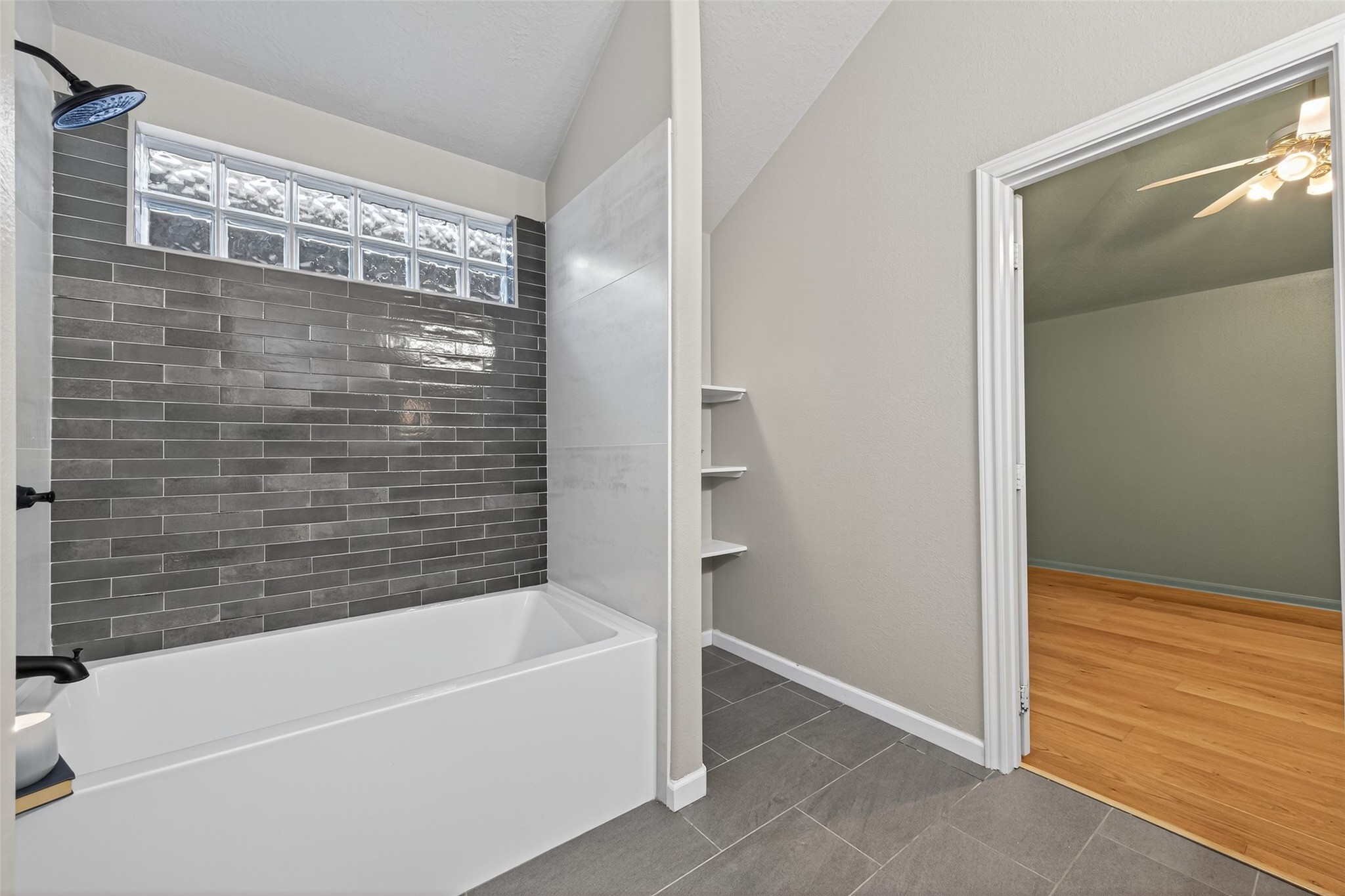 2910 Crescent Bend Road Spring, TX 77388 - Photo 15 of 29 This bathroom features a modern design with a sleek bathtub against a gray tiled wall, glass block window for natural light, and an adjacent room with wooden flooring and a ceiling fan.