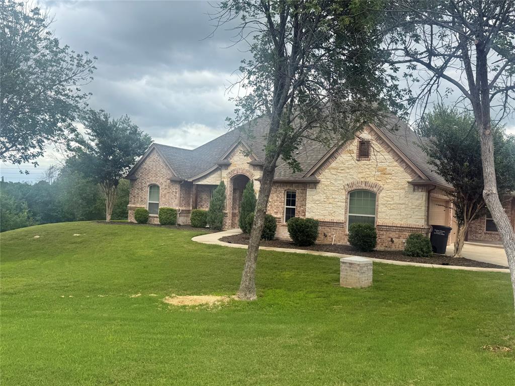 101 Bedding Lane Azle, TX 76020 - Photo 2 of 6 Exterior featuring a blend of brick and stone masonry, a dark shingle roof, a curving concrete walkway, manicured shrubs, and mature trees
