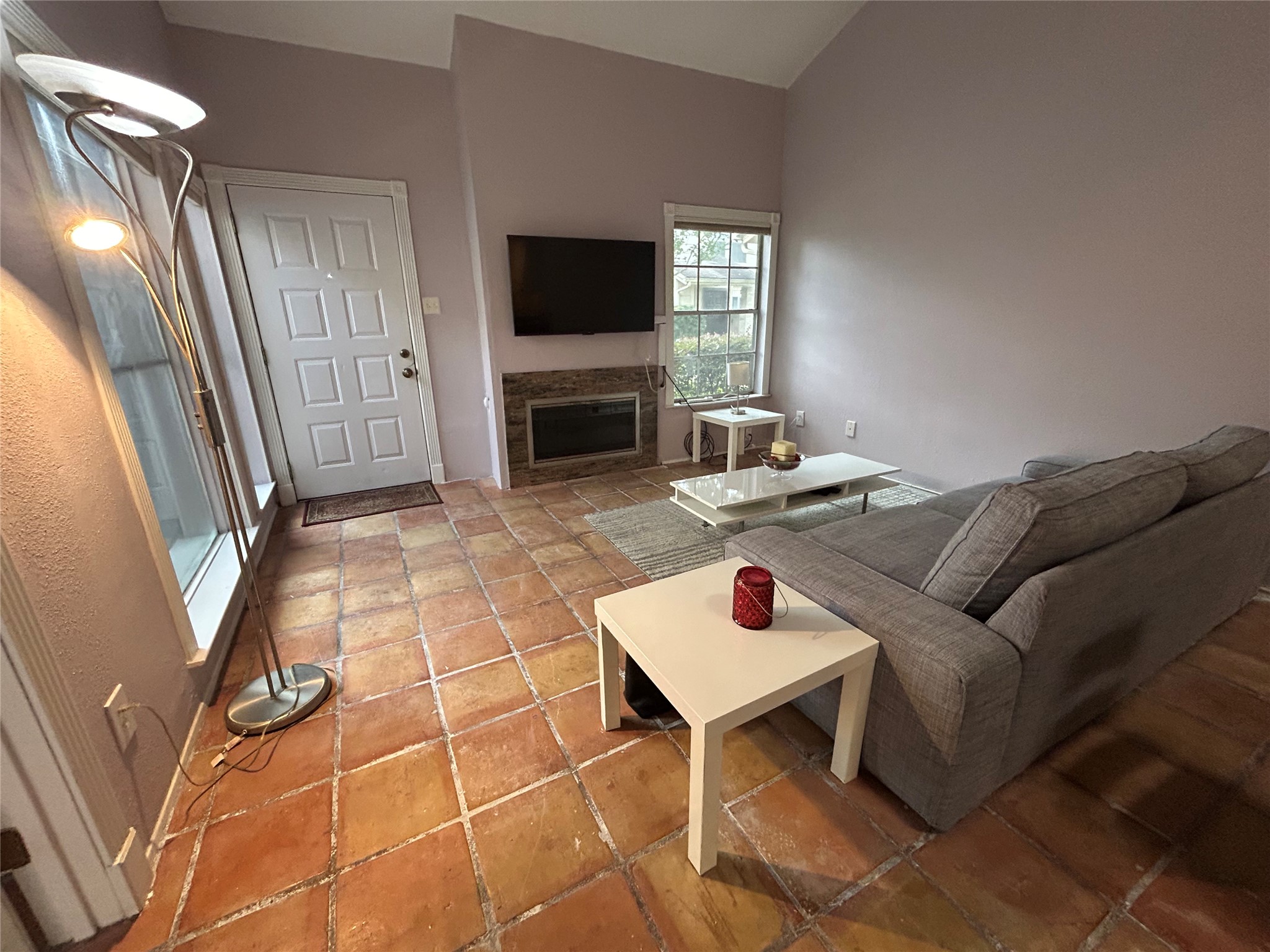 13034 Leader Street, Unit 965 Houston, TX 77072 - Photo 1 of 32 This cozy living room features terracotta tile flooring, a mounted TV above a fireplace, and large windows for natural light. The space is furnished with a modern gray couch and sleek coffee table, creating a welcoming atmosphere.