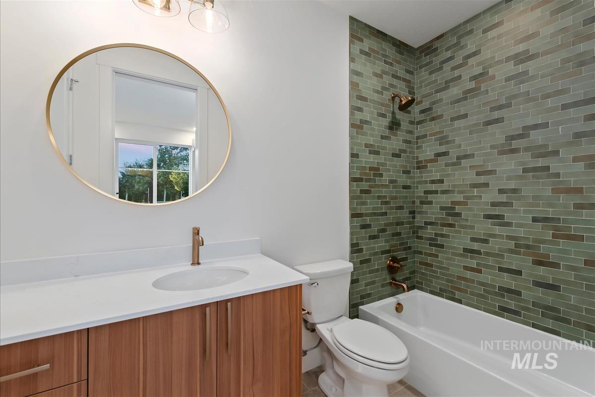 3213 West Moore Street Boise, ID 83702 - Photo 27 of 32 Bathroom featuring a wood-finish vanity with a white countertop and undermount sink, a circular mirror with a gold-tone frame, green subway tile shower surround, and bronze-tone fixtures