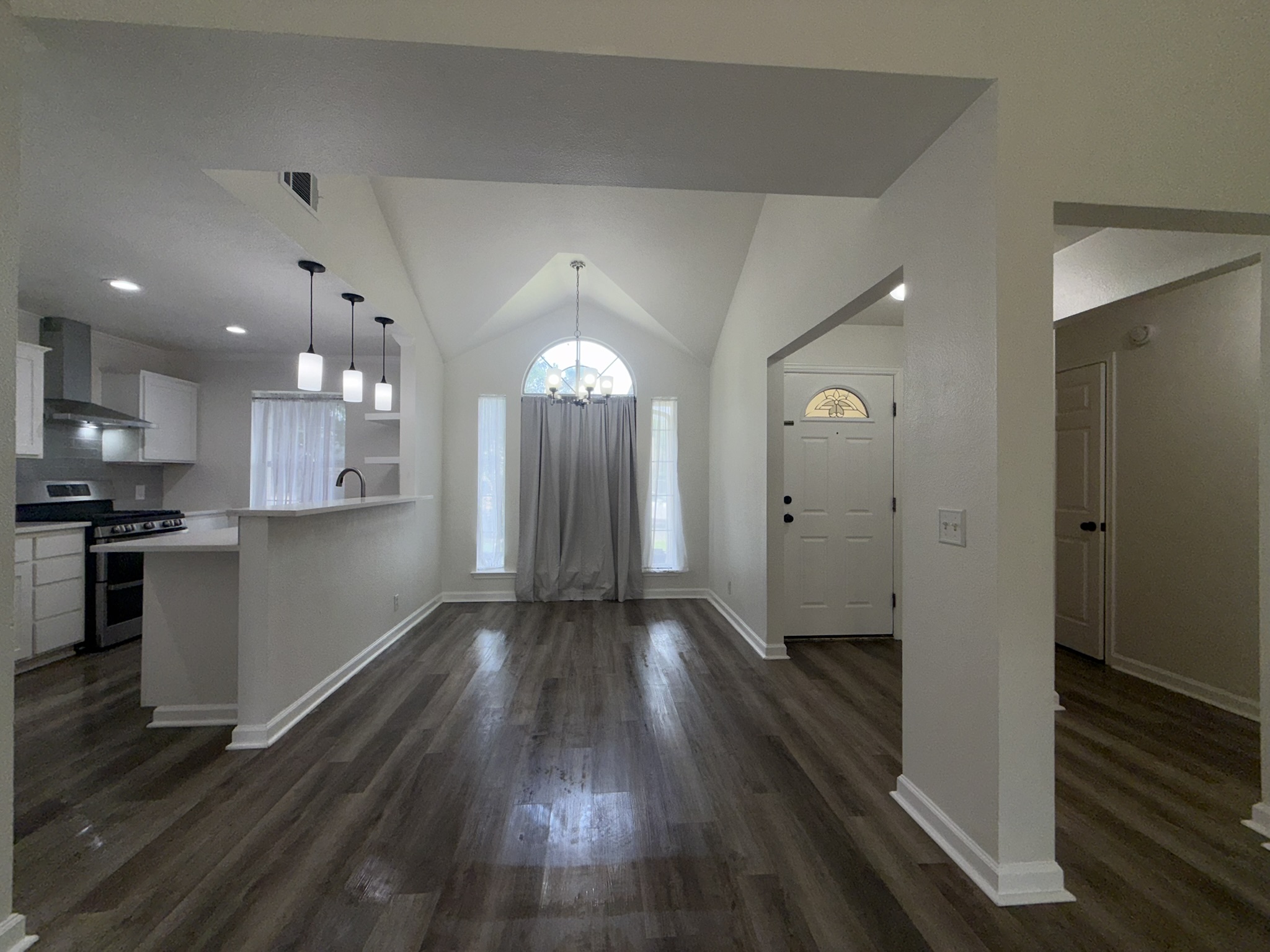 Expansive open concept living area featuring vaulted ceilings, wood-finish flooring, a kitchen island with pendant lighting, a built-in oven, and a decorative front door with an arched transom window
