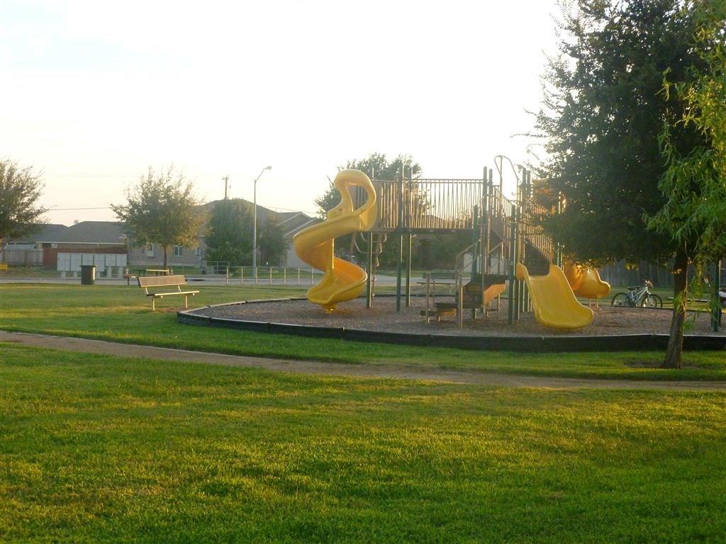 107 Inman Drive Hutto, TX 78634 - Photo 18 of 19 Expansive green space featuring a playground with yellow slides, a climbing structure, a concrete pathway, a wooden park bench, and mature trees