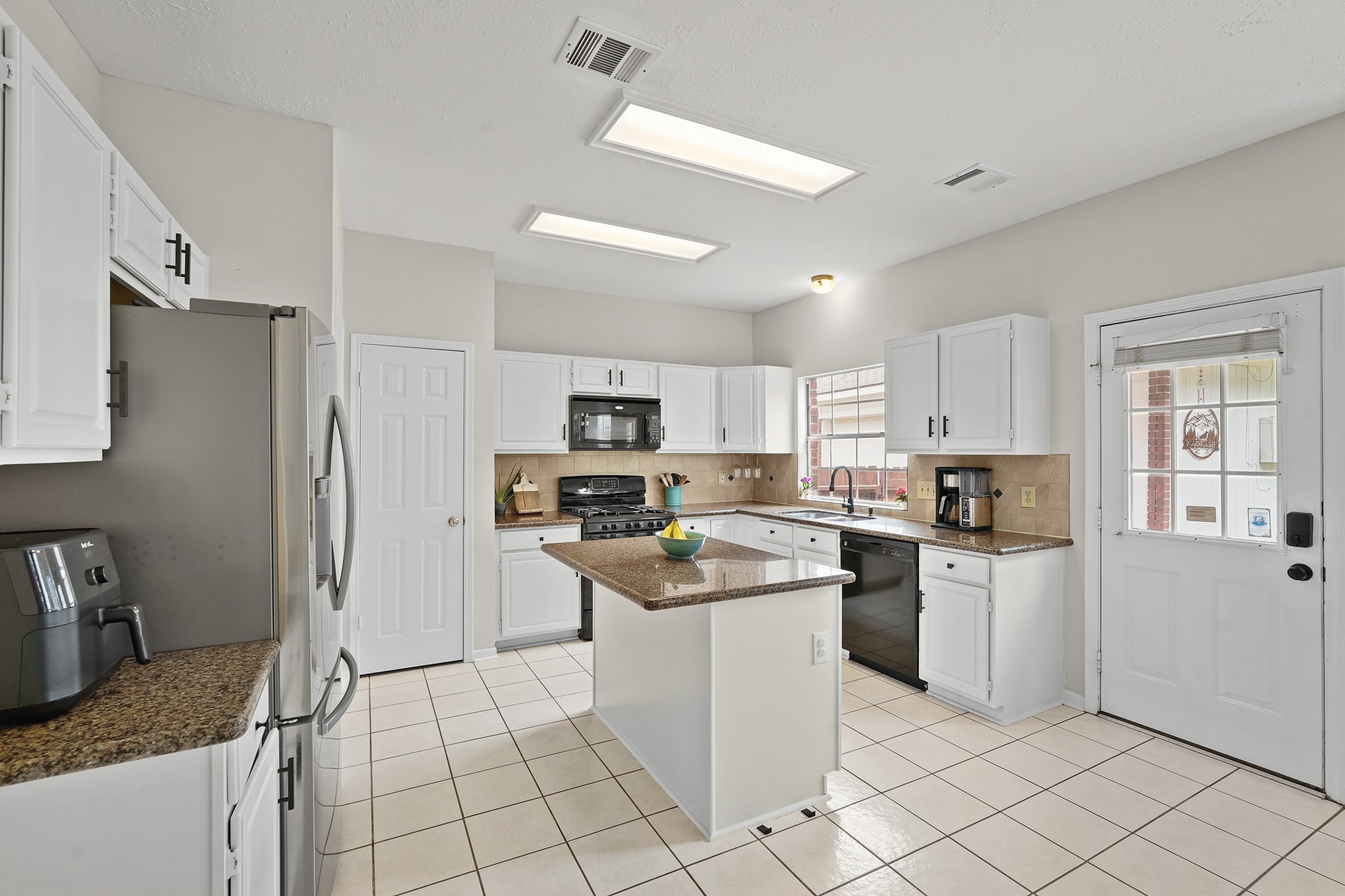 16114 Kintyre Point Road Houston, TX 77095 - Photo 19 of 44 This kitchen features a clean and bright design with white cabinets and tiled flooring. It includes modern appliances, an island with granite countertops, and ample natural light from a window above the sink and a glass-paneled door. Perfect for a functional and inviting cooking space.