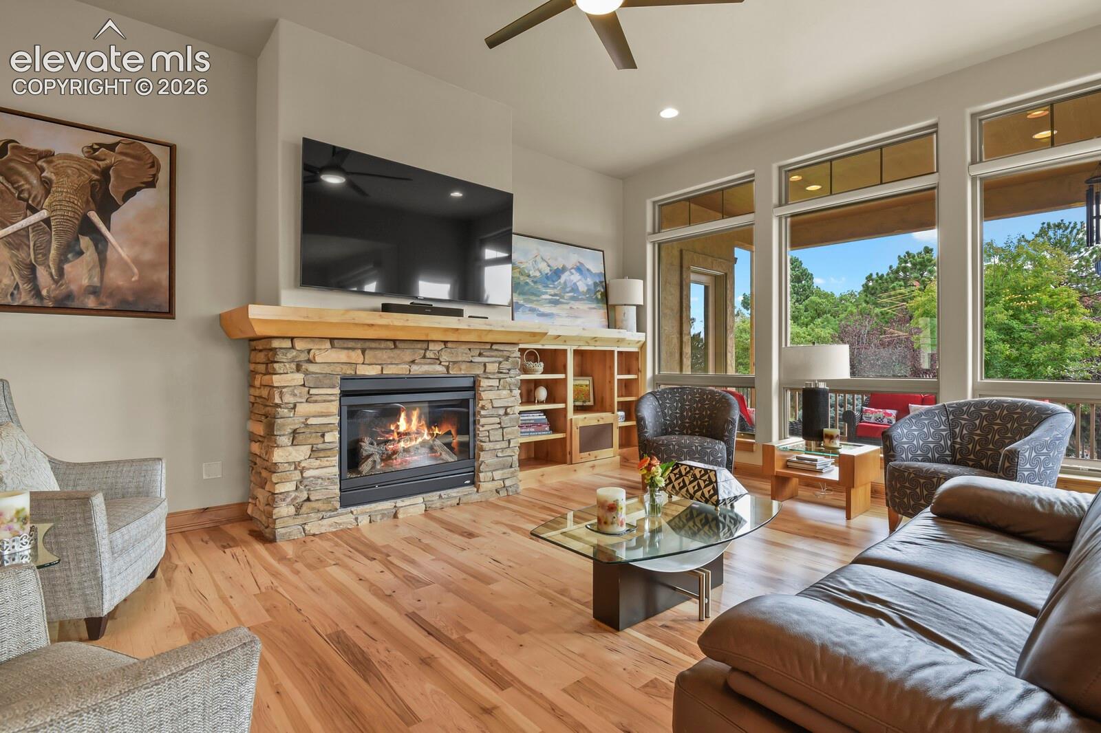 Stone-clad fireplace with wood mantel, custom hardwood flooring, built-in shelving, expansive windows, and a ceiling fan
