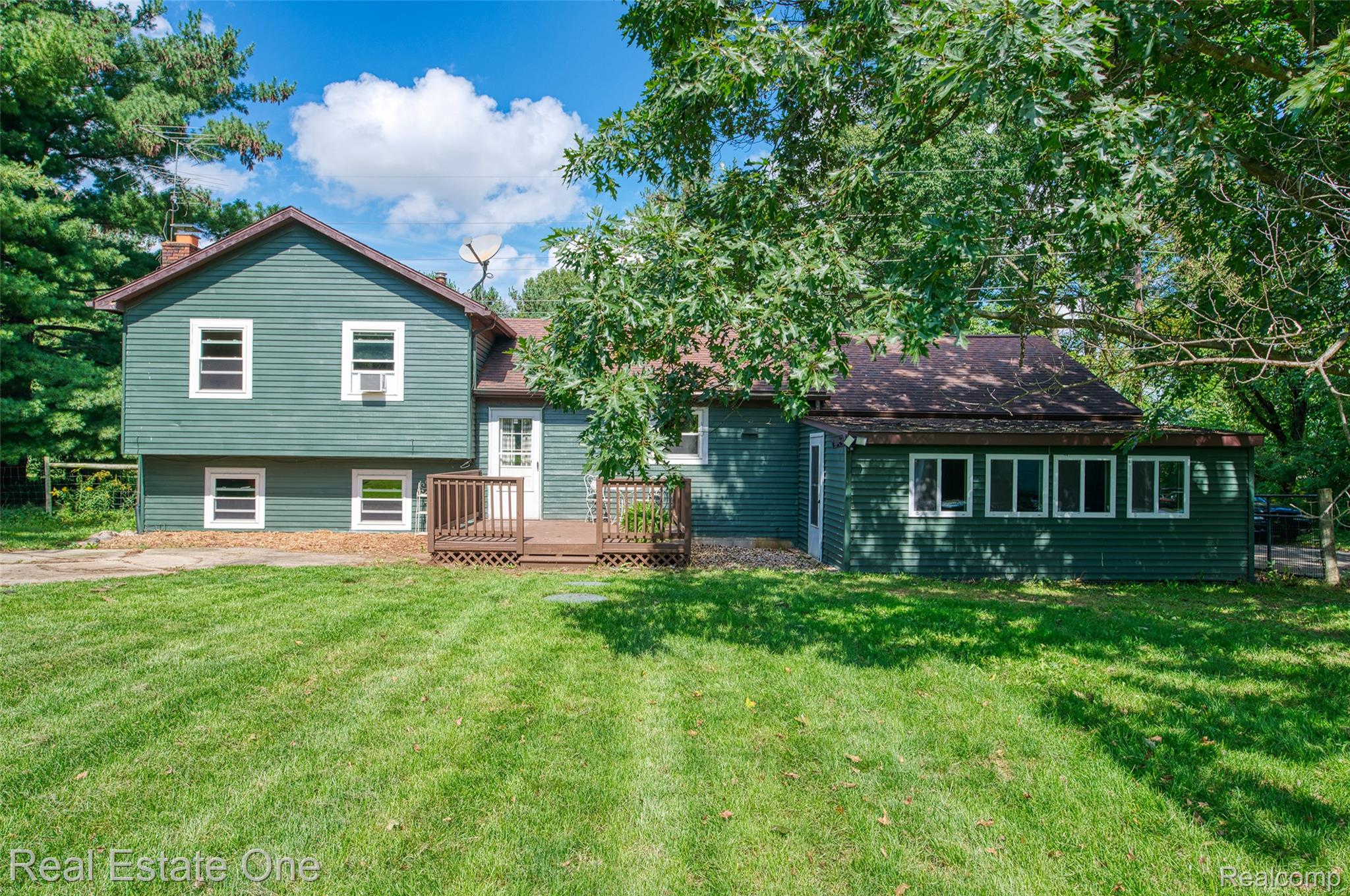 5130 Iosco Road Webberville, MI 48892 - Photo 6 of 39 Expansive green lawn with mature trees, a two-story exterior in a dark green finish, a wood-finish deck, and a sunroom with multiple windows