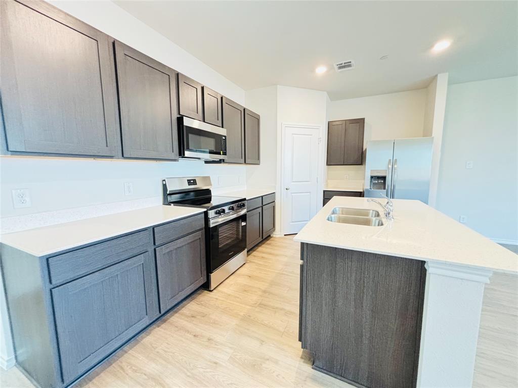 120 Redwood Road Princeton, TX 75407 - Photo 4 of 24 Modern kitchen featuring wood-finish flooring, white countertops, dark wood-finish cabinetry, stainless steel appliances, and a central island with a double basin sink.