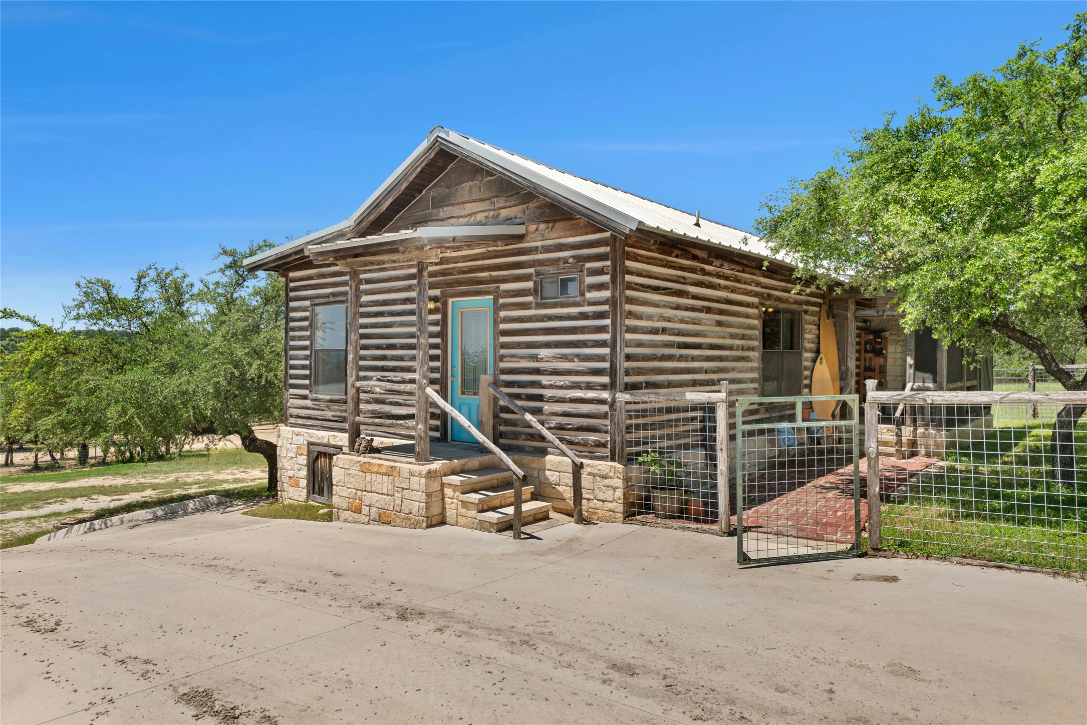 496 Middleton Road Spicewood, TX 78669 - Photo 4 of 40 Log cabin exterior featuring a turquoise entry door, stone foundation, covered porch, metal roof, and a fenced patio area