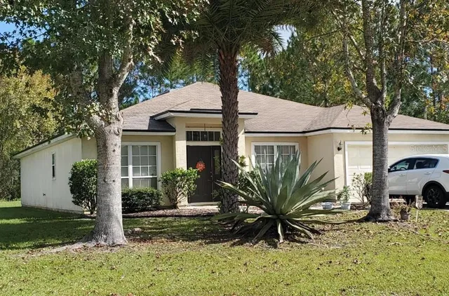 a view of a white house next to a yard with potted plants and large trees