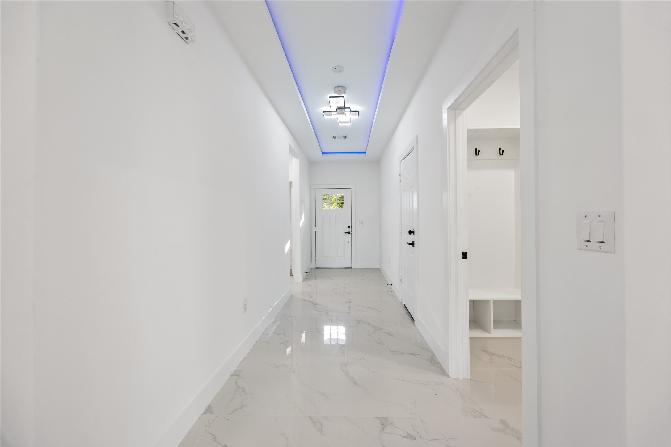 1121 Austin Street Rosenberg, TX 77471 - Photo 13 of 44 This photo shows a bright, modern hallway with glossy white large format porcelain floors and sleek white walls. The ceiling features a stylish light fixture with blue accent lighting. A door at the end leads outside, and there's a small nook with hooks and cubbies on the right.