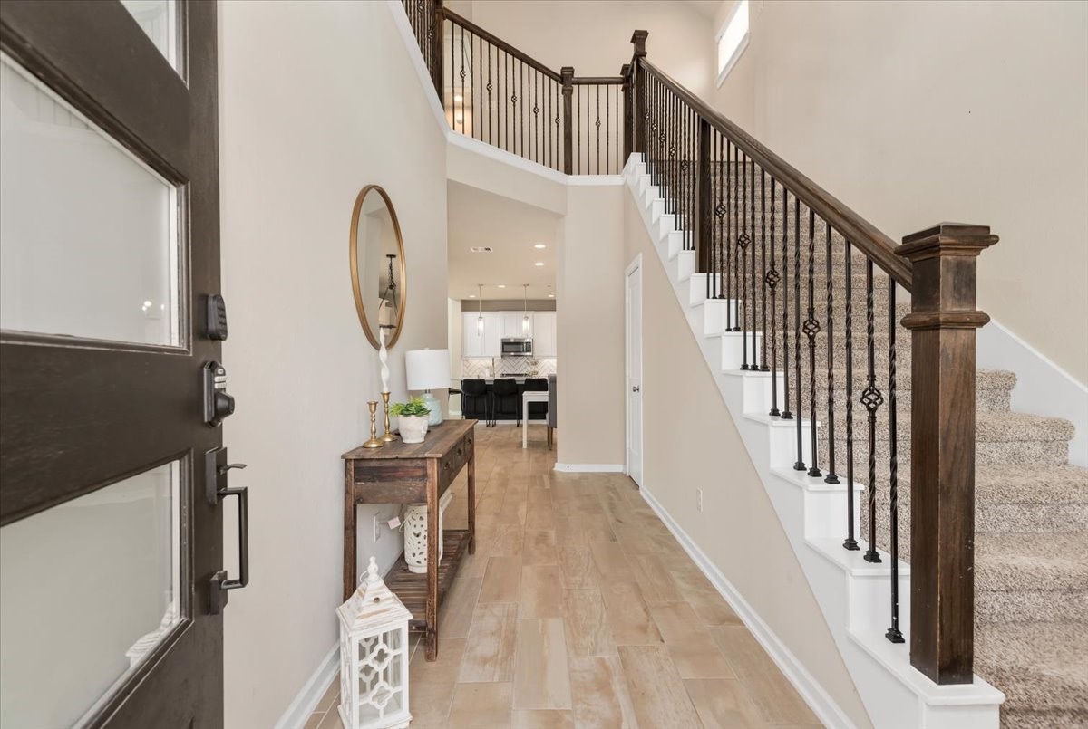 Bright and inviting entryway featuring soaring ceilings, neutral tones, and durable tile flooring throughout the main walkway. Elegant staircase with wrought iron balusters and rich wood handrails creates a striking first impression. The open-concept layout offers a seamless view into the living and kitchen areas, enhancing the home’s spacious feel. A stylish console table adds a welcoming touch, while abundant natural light highlights the clean, modern finishes.