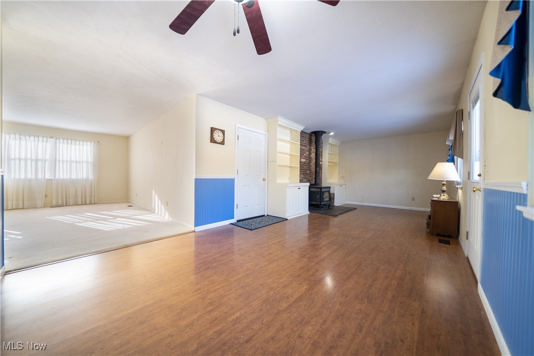 Unfurnished family room featuring a wood stove, dark wood finished floors, a wainscoted wall, and ceiling fan
