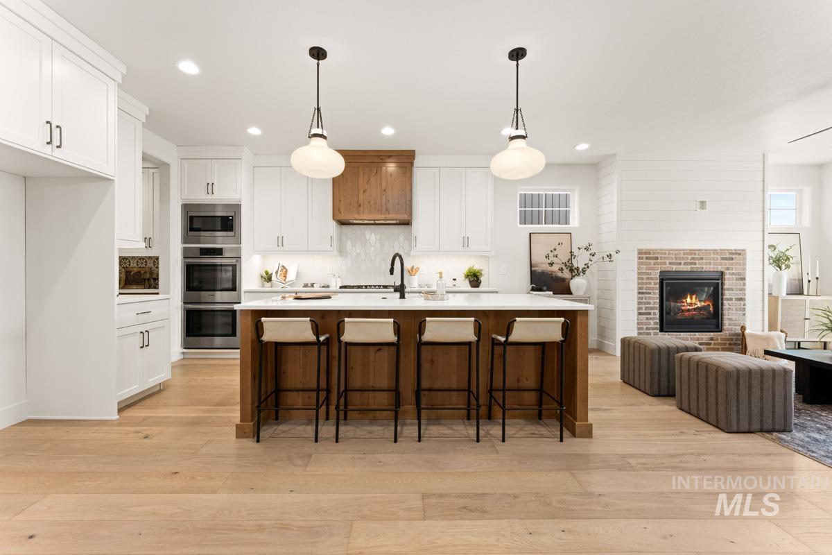 13338 North Spring Creek Way Boise, ID 83714 - Photo 8 of 46 Open-concept kitchen and living area featuring light wood-finish flooring, a kitchen island with a light-toned countertop, white cabinetry, integrated stainless steel appliances, and a brick fireplace