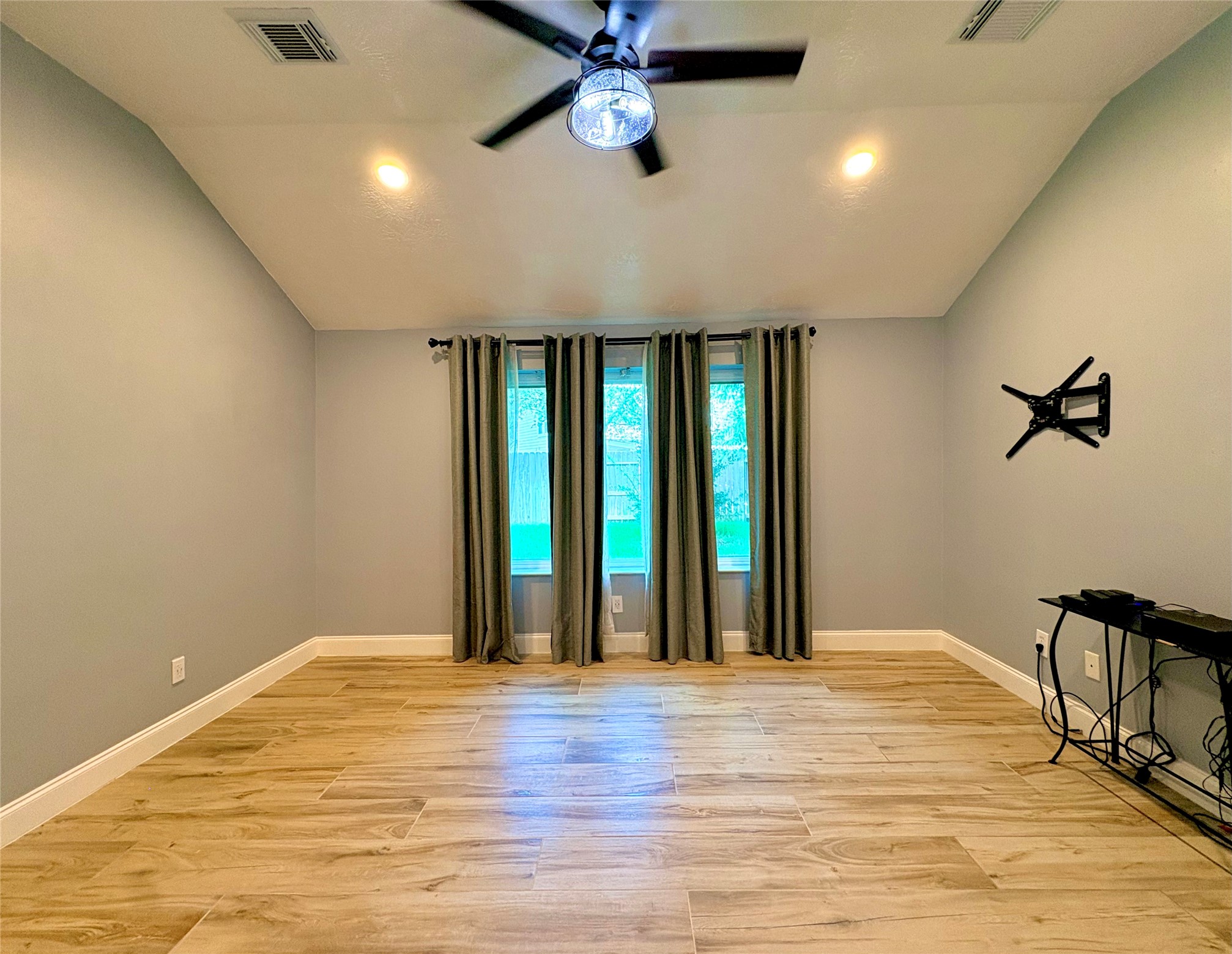 15410 Redbud Berry Way Cypress, TX 77433 - Photo 20 of 49 Primary room features a spacious area with light wood flooring, soft gray walls, and a ceiling fan. It has a large window with curtains, providing natural light, and a wall mount ready for a TV.
