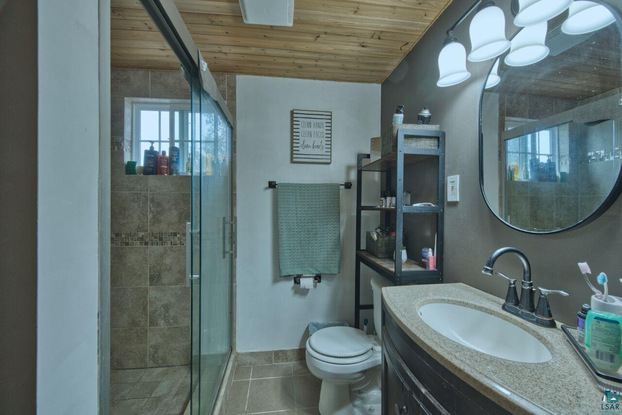 329 Wyandotte Road Hoyt Lakes, MN 55750 - Photo 21 of 52 Bathroom featuring a glass-enclosed shower with tile surround, wood-finish ceiling, stone-look countertop, dark cabinetry, and a circular wall mirror