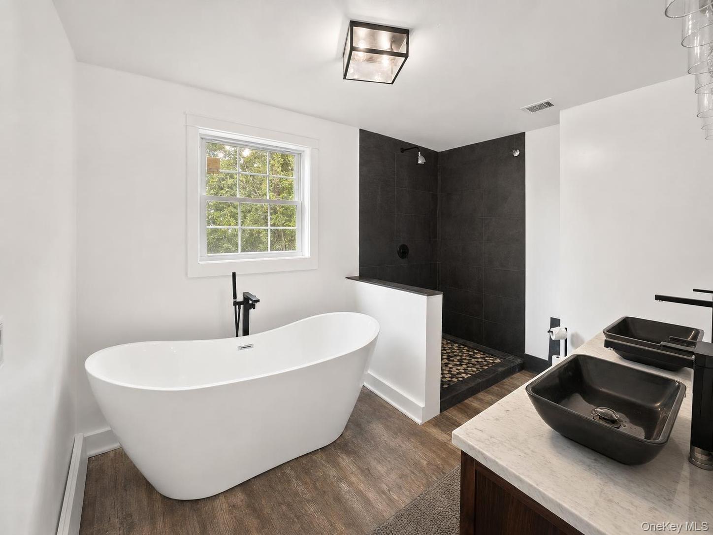 150 Cold Spring Road Liberty, NY 12754 - Photo 11 of 25 Freestanding soaking tub, wood-finish flooring, walk-in shower with dark tile surround and mosaic floor, vanity with two vessel sinks, and dark fixtures