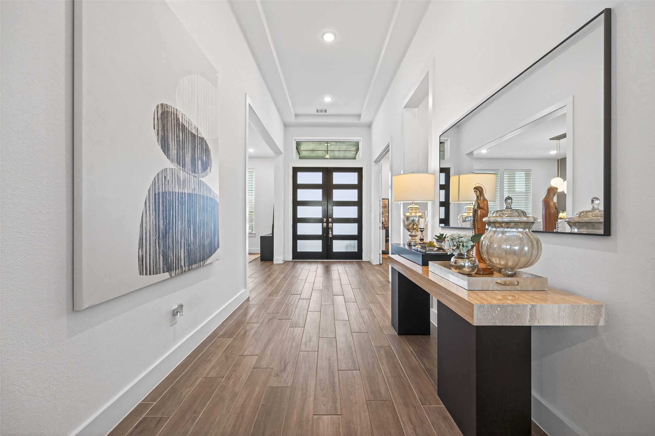 29612 Fulshear Lake Trace Fulshear, TX 77406 - Photo 12 of 48 This photo showcases a modern, well-lit hallway with wooden flooring and contemporary decor. It features a stylish front door with frosted glass panels and tasteful artwork on the walls. A sleek console table with decorative items and a large mirror enhance the elegance of the space.