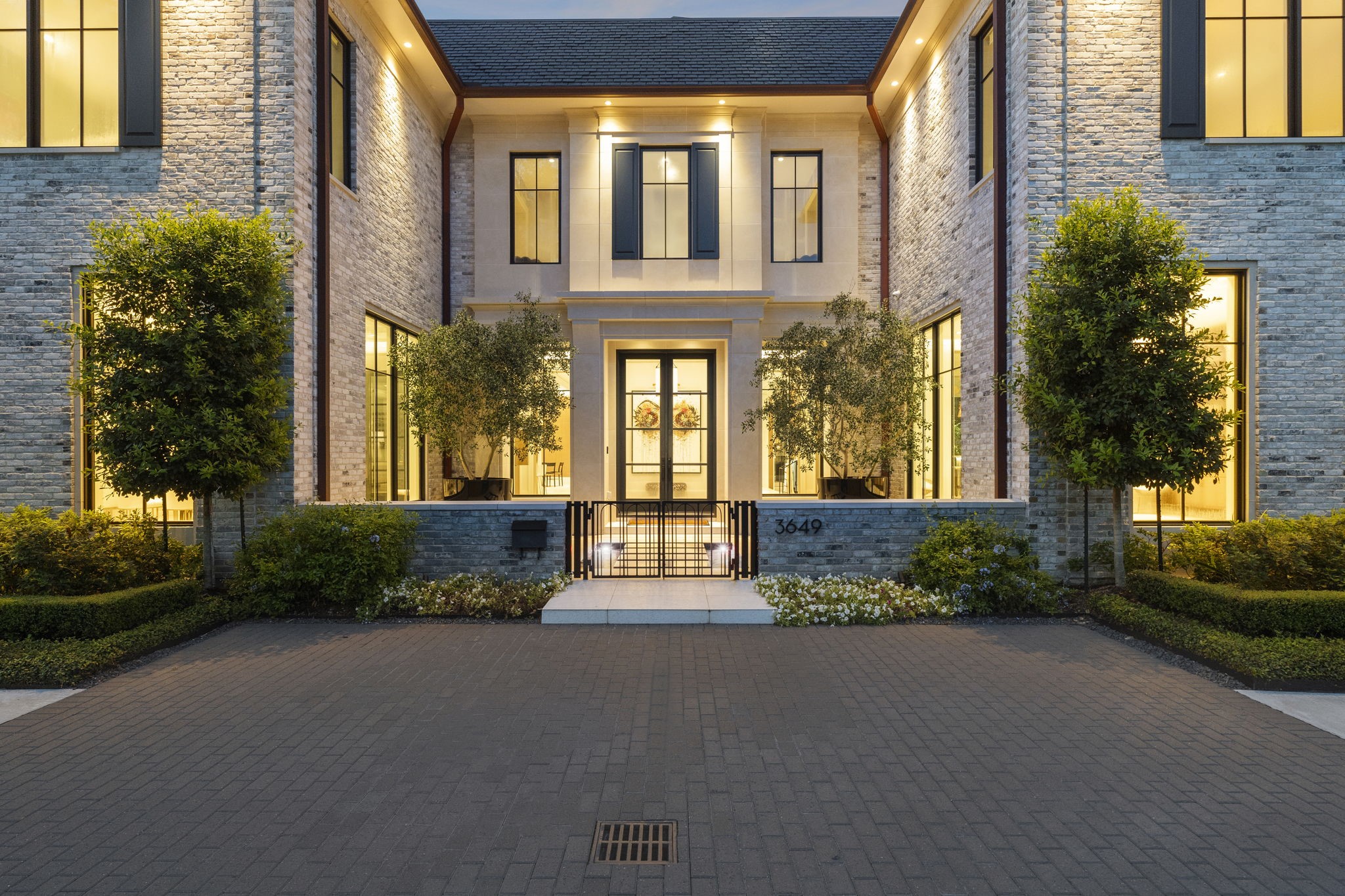 Striking front elevation with classic brick exterior, oversized windows, and a gated motor court. Manicured landscaping and ambient lighting frame the elegant glass entry, setting a refined tone upon arrival.