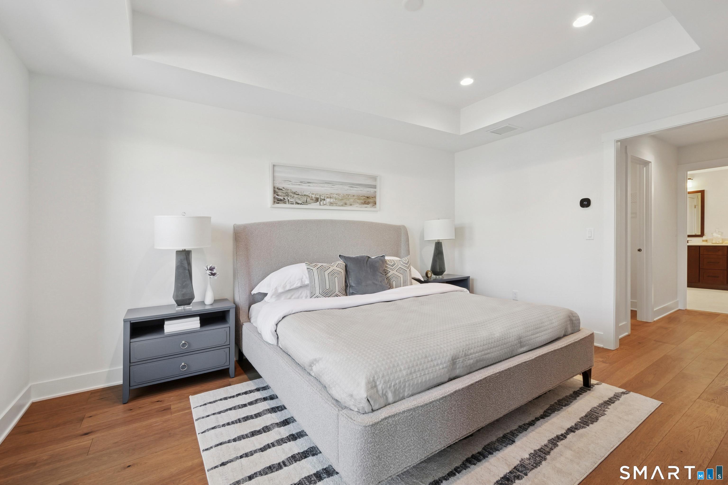 5 Edgewood Way, Unit 25 Woodbridge, CT 06525 - Photo 3 of 8 staged model Silliman Modern Farmhouse showing primary bedroom with king-size bed and two large end tables. Down the hall is the walk-in closet and ensuite bath with double vanity and expanded seated shower