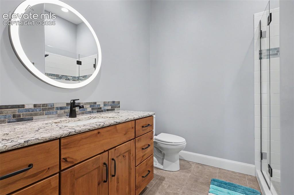 21 Leaming Road Colorado Springs, CO 80906 - Photo 35 of 48 Bathroom featuring a light wood-finish vanity with a stone countertop, a tiled backsplash, and a round illuminated mirror