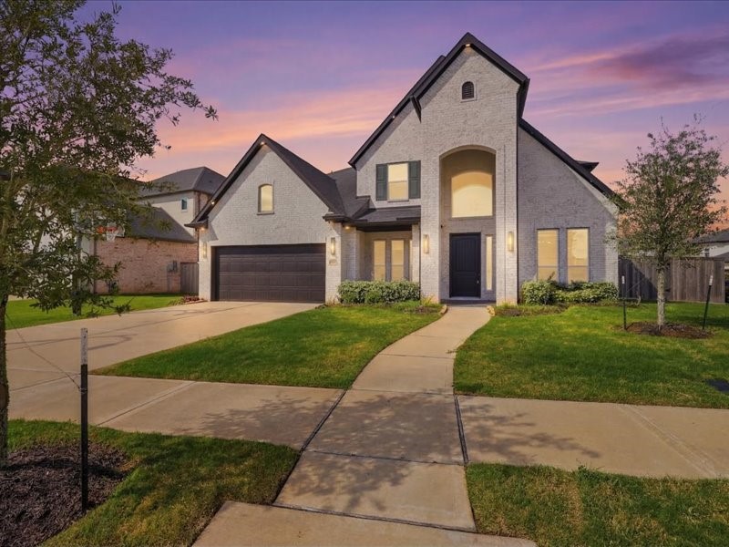 This photo showcases a modern two-story home with a well-maintained front lawn, a spacious driveway leading to a two-car garage, and a welcoming entrance. The exterior features a mix of brick and siding, set against a picturesque sunset backdrop.