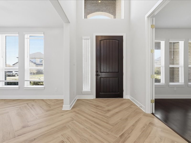 4710 Pecos Ridge Lane Sugar Land, TX 77479 - Photo 13 of 49 This photo shows a bright, welcoming entryway with high ceilings and abundant natural light. It features a dark wooden front door, light wood herringbone flooring, and large windows with white blinds, giving it a modern and airy feel. A room with dark flooring is visible to the right.