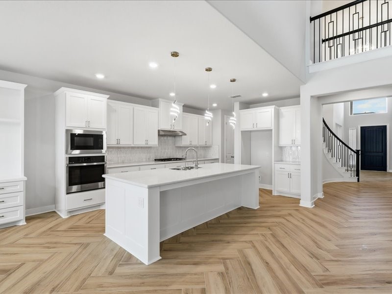 4710 Pecos Ridge Lane Sugar Land, TX 77479 - Photo 15 of 49 This modern kitchen features a spacious open layout with sleek white cabinetry, a large center island, stainless steel appliances, and elegant pendant lighting. The herringbone wood flooring adds warmth, while the nearby staircase and high ceilings create an airy, inviting atmosphere.