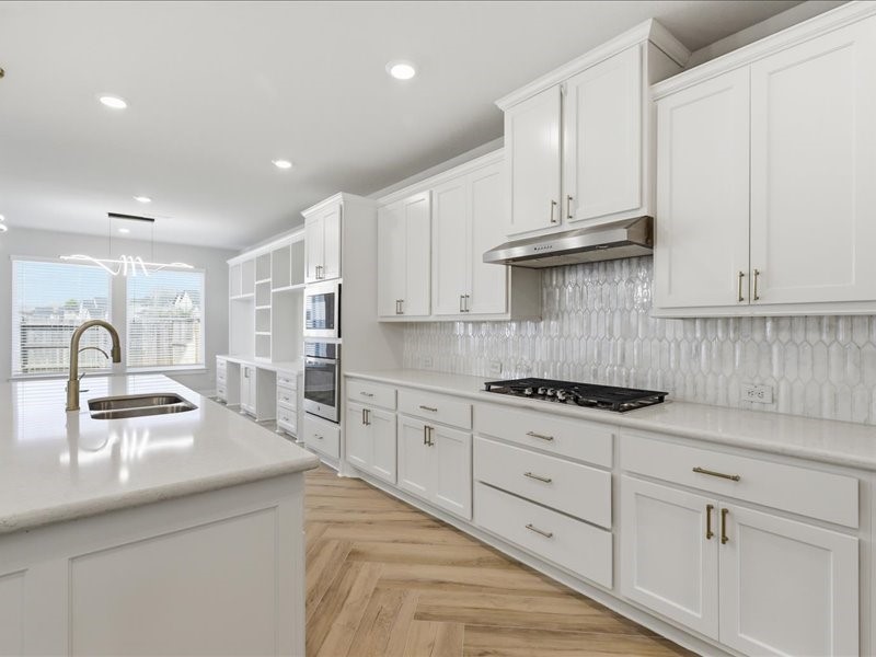 4710 Pecos Ridge Lane Sugar Land, TX 77479 - Photo 18 of 49 This spacious kitchen features sleek white cabinetry, modern stainless steel appliances, and a stylish tile backsplash. It boasts a large island with a sink, elegant gold fixtures, and ample natural light from large windows, complemented by recessed lighting. Perfect for both cooking and entertaining.