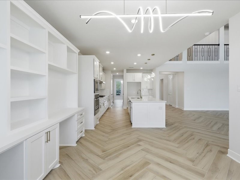 4710 Pecos Ridge Lane Sugar Land, TX 77479 - Photo 23 of 49 This photo showcases a modern, open-concept kitchen with sleek white cabinetry, a central island, and integrated appliances. The space features elegant herringbone wood flooring and a distinctive contemporary light fixture, with high ceilings and a view into adjoining rooms.