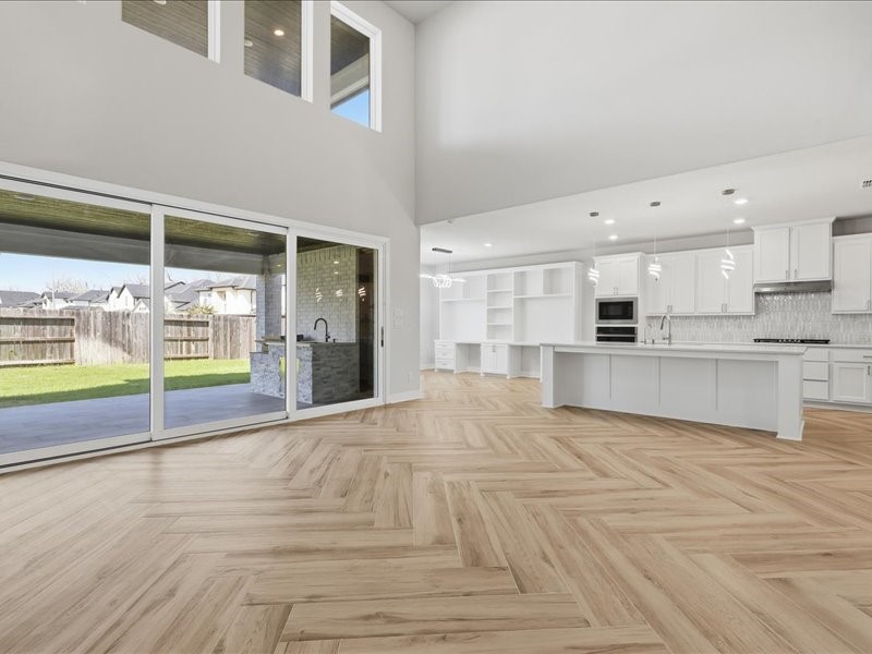 4710 Pecos Ridge Lane Sugar Land, TX 77479 - Photo 26 of 49 This spacious, modern open-plan living area features high ceilings, abundant natural light, and elegant light wood herringbone flooring. It includes a sleek white kitchen with an island, and large sliding glass doors leading to a covered outdoor patio and grassy backyard.