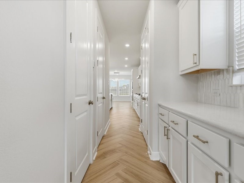 4710 Pecos Ridge Lane Sugar Land, TX 77479 - Photo 30 of 49 A bright, narrow hallway with sleek white cabinetry on the right and elegant herringbone wood flooring. The space leads to an open room with large windows, allowing natural light to fill the area.