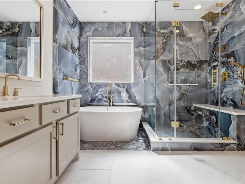 4710 Pecos Ridge Lane Sugar Land, TX 77479 - Photo 36 of 49 This modern bathroom features a luxurious freestanding tub, a spacious glass-enclosed shower with gold fixtures, and stunning marble-patterned walls. The vanity offers ample storage with sleek cabinetry, and the room is filled with natural light from a large window.