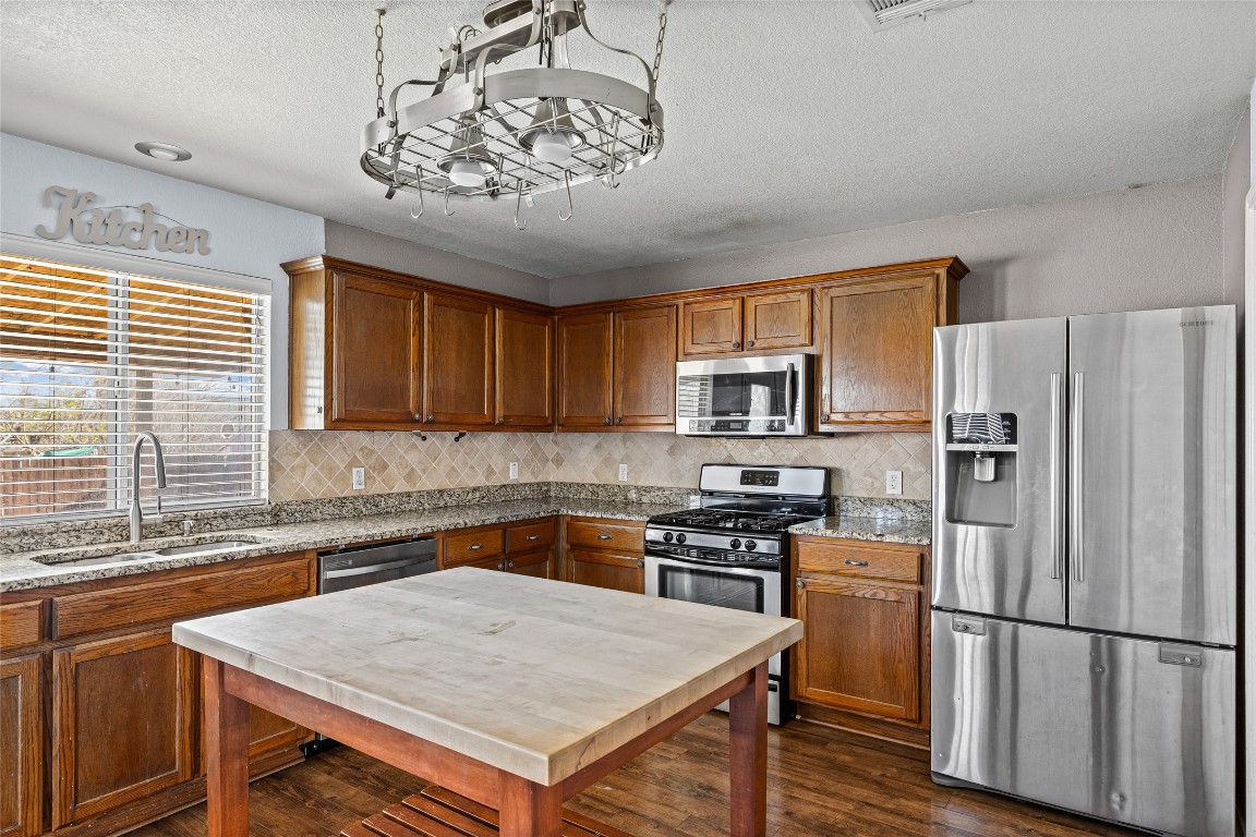 1321 Bunratty Circle Pflugerville, TX 78660 - Photo 16 of 31 Kitchen featuring stainless steel appliances, brown cabinets, light stone countertops, dark wood-type flooring, and a textured ceiling