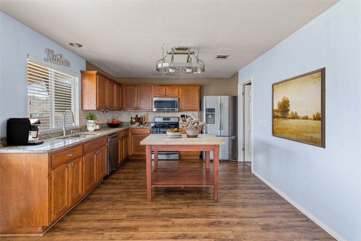 1321 Bunratty Circle Pflugerville, TX 78660 - Photo 8 of 31 Kitchen featuring brown cabinetry, appliances with stainless steel finishes, dark wood-type flooring, and light stone countertops