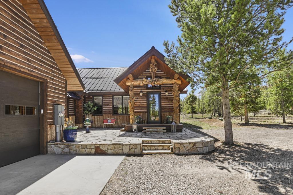65 Goat Creek Way Stanley, ID 83278 - Photo 2 of 32 Log cabin exterior with a dark metal roof, stone patio, and a two-car garage featuring a dark wood-finish door