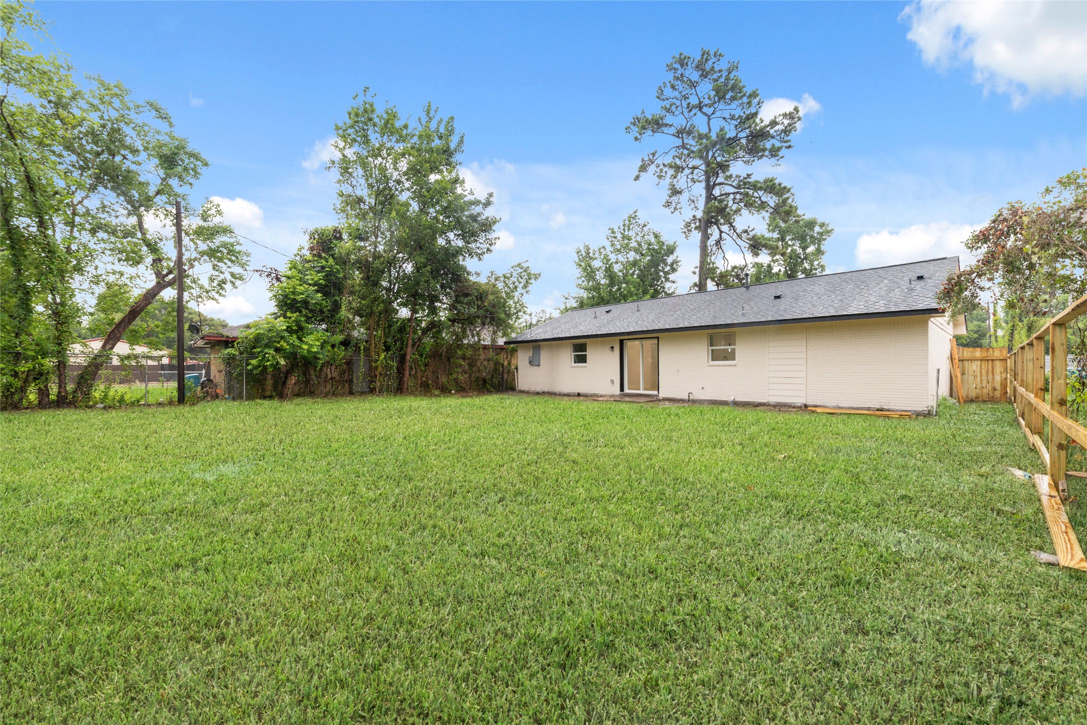 5709 Langley Road Houston, TX 77016 - Photo 25 of 27 This photo shows a spacious backyard with a well-maintained lawn, surrounded by a wooden fence and mature trees. The back of the house is visible, featuring a light exterior and multiple windows, offering a private and inviting outdoor space.