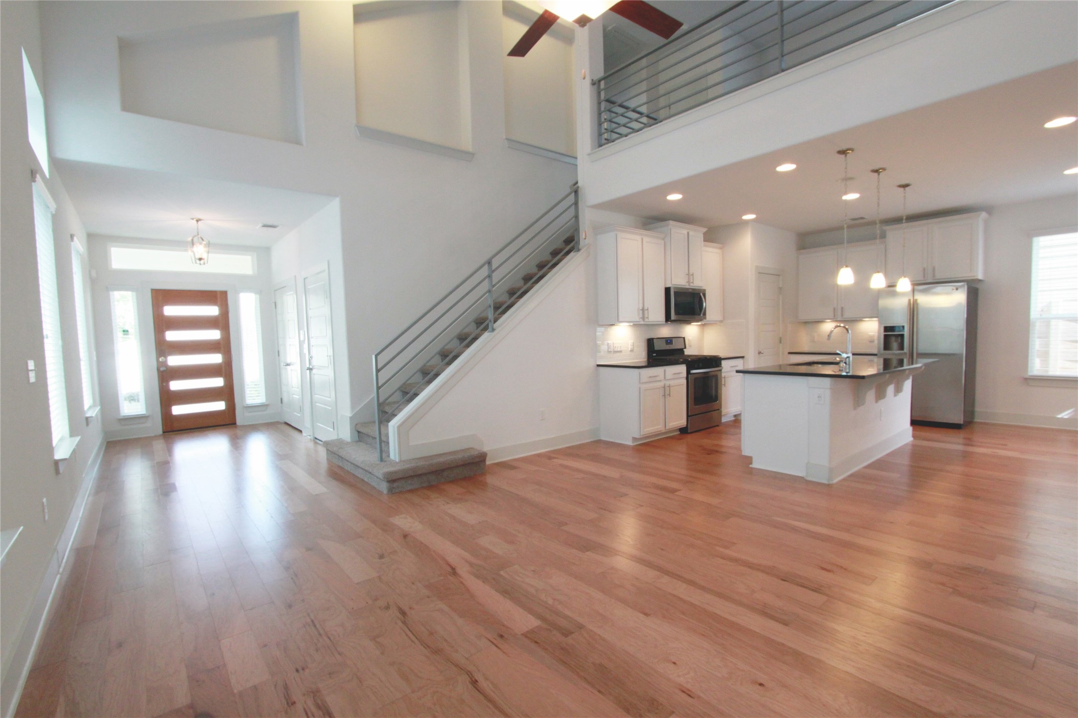 1507 Homespun Road Austin, TX 78745 - Photo 8 of 35 Open-concept living space featuring wood-finish flooring, a contemporary kitchen with a central island and white cabinetry, a prominent staircase with metal railing, a two-story ceiling, and a modern entry door with sidelights