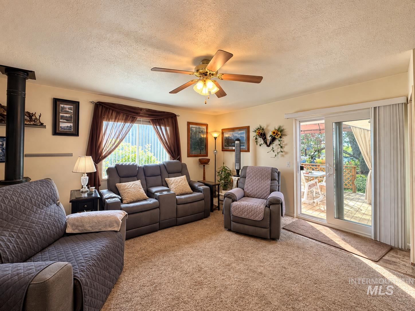 181 Hillcrest Road Burley, ID 83318 - Photo 6 of 14 Carpeted living area featuring a wood-burning stove, ceiling fan, large window, and sliding glass door leading to an outdoor patio