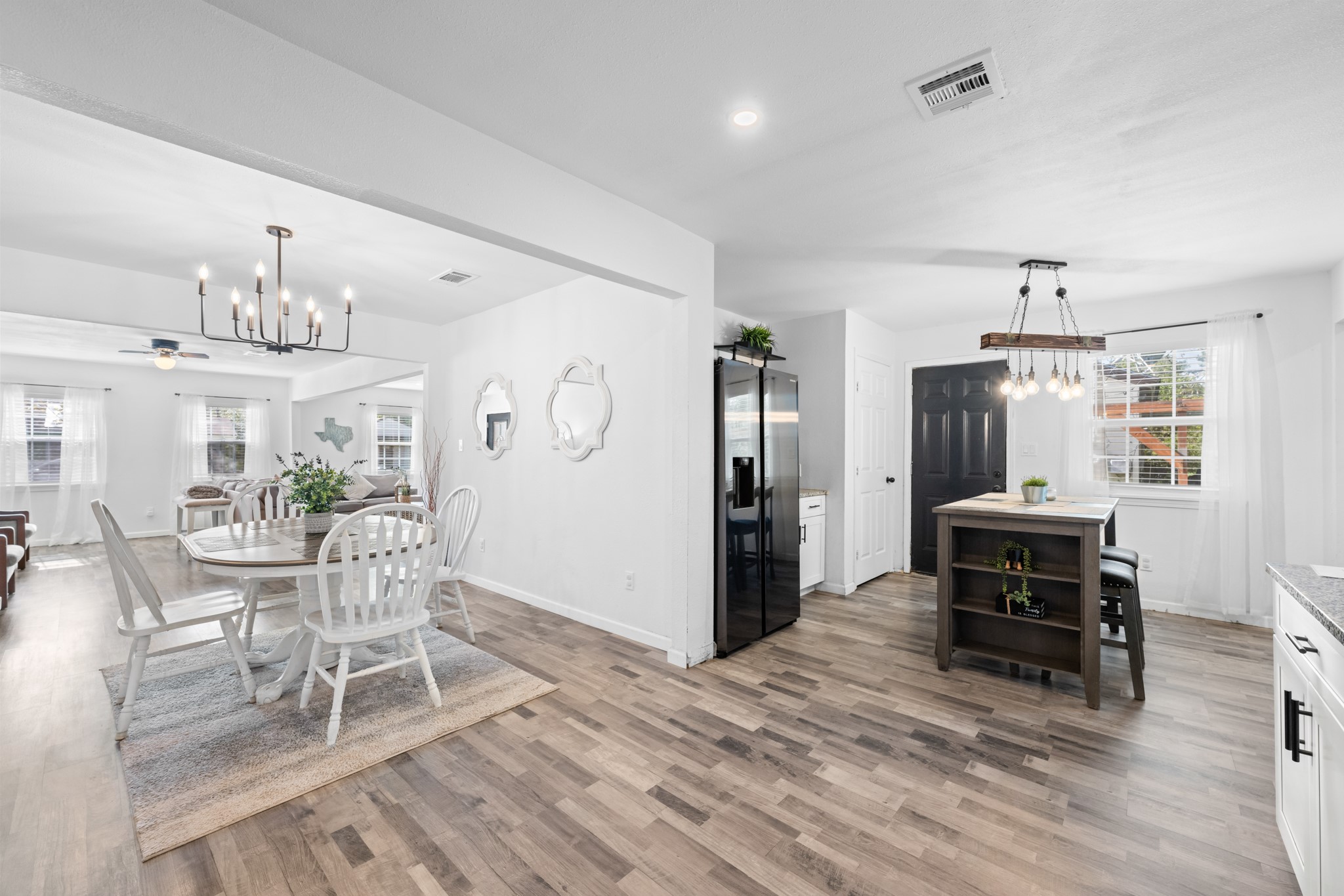 729 Kyle Angleton, TX 77515 - Photo 12 of 36 This photo showcases a bright, open-concept dining and kitchen area featuring modern lighting, white walls, and wood-look flooring. The dining area has a round table with white chairs, while the kitchen includes a central island and large windows for natural light.